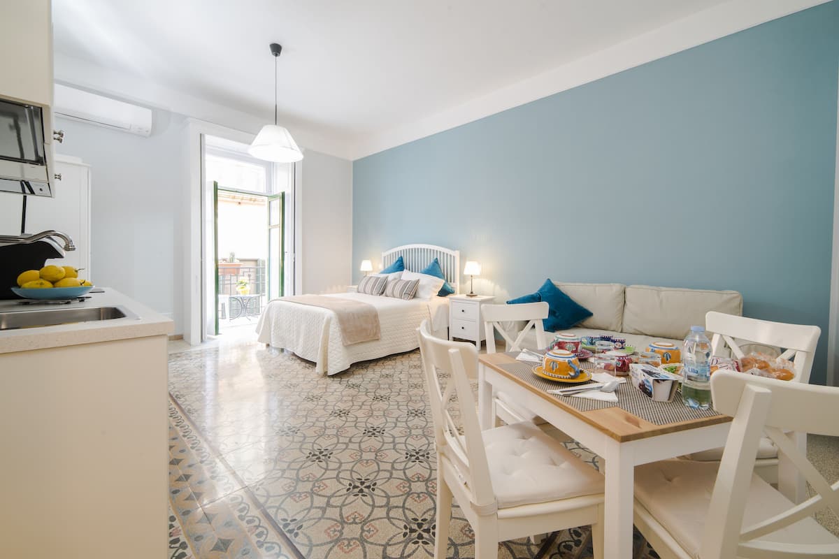 Quarto de hotel moderno com cozinha compacta, cama com cabeceira branca, área de jantar com mesa e cadeiras, e uma parede de destaque azul.