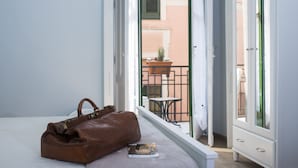 Un lit avec un sac en cuir marron et un livre, une fenêtre donnant sur un balcon et du mobilier d’extérieur.