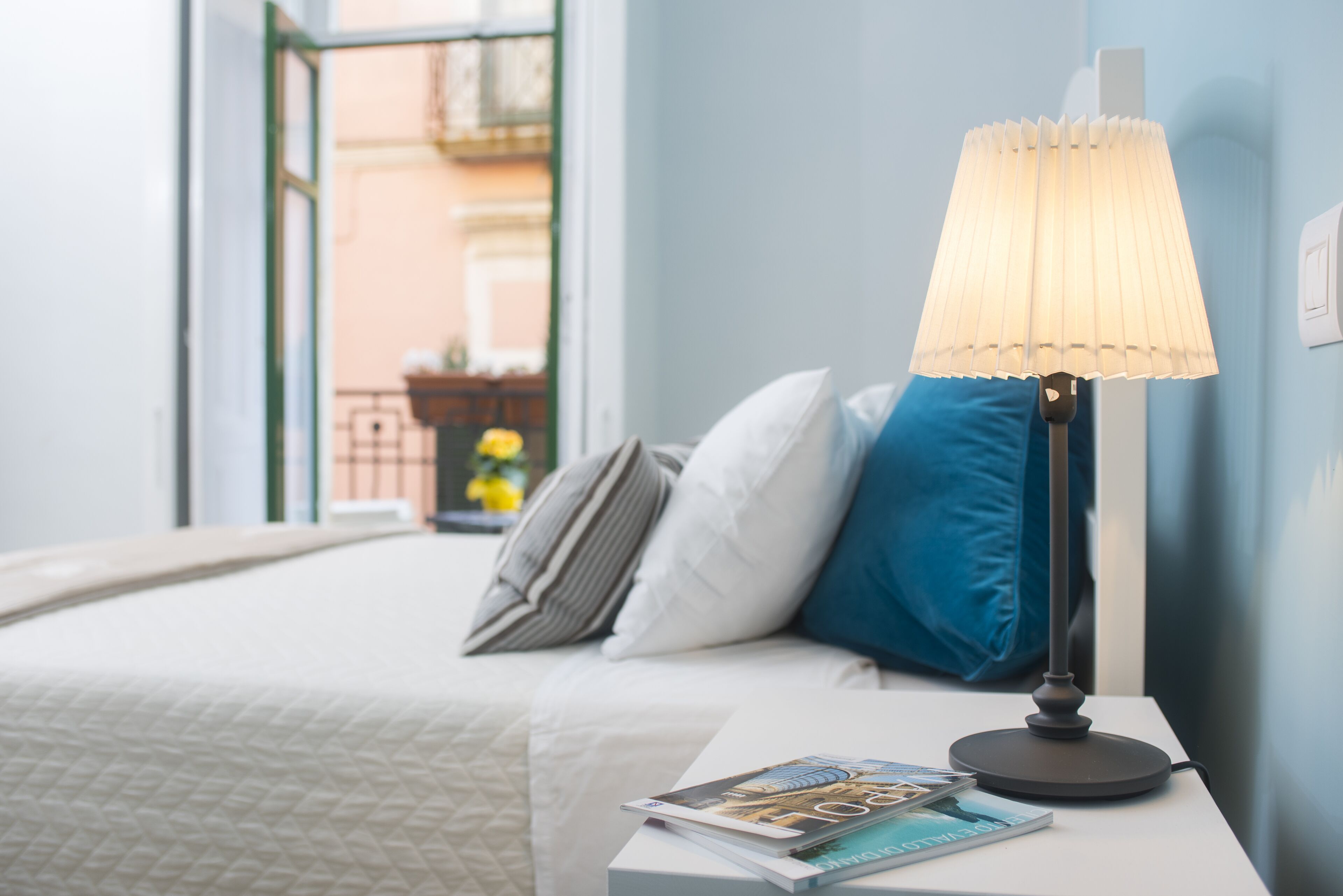 A neatly made bed with a white duvet and pillows, a bedside table with a lamp, and a magazine.