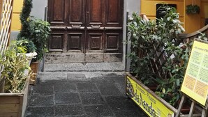 Une grande porte en bois richement décorée, surmontée d’une arche ornementale, est encadrée dans le mur d’un bâtiment jaune, devant lequel se trouve une enseigne.