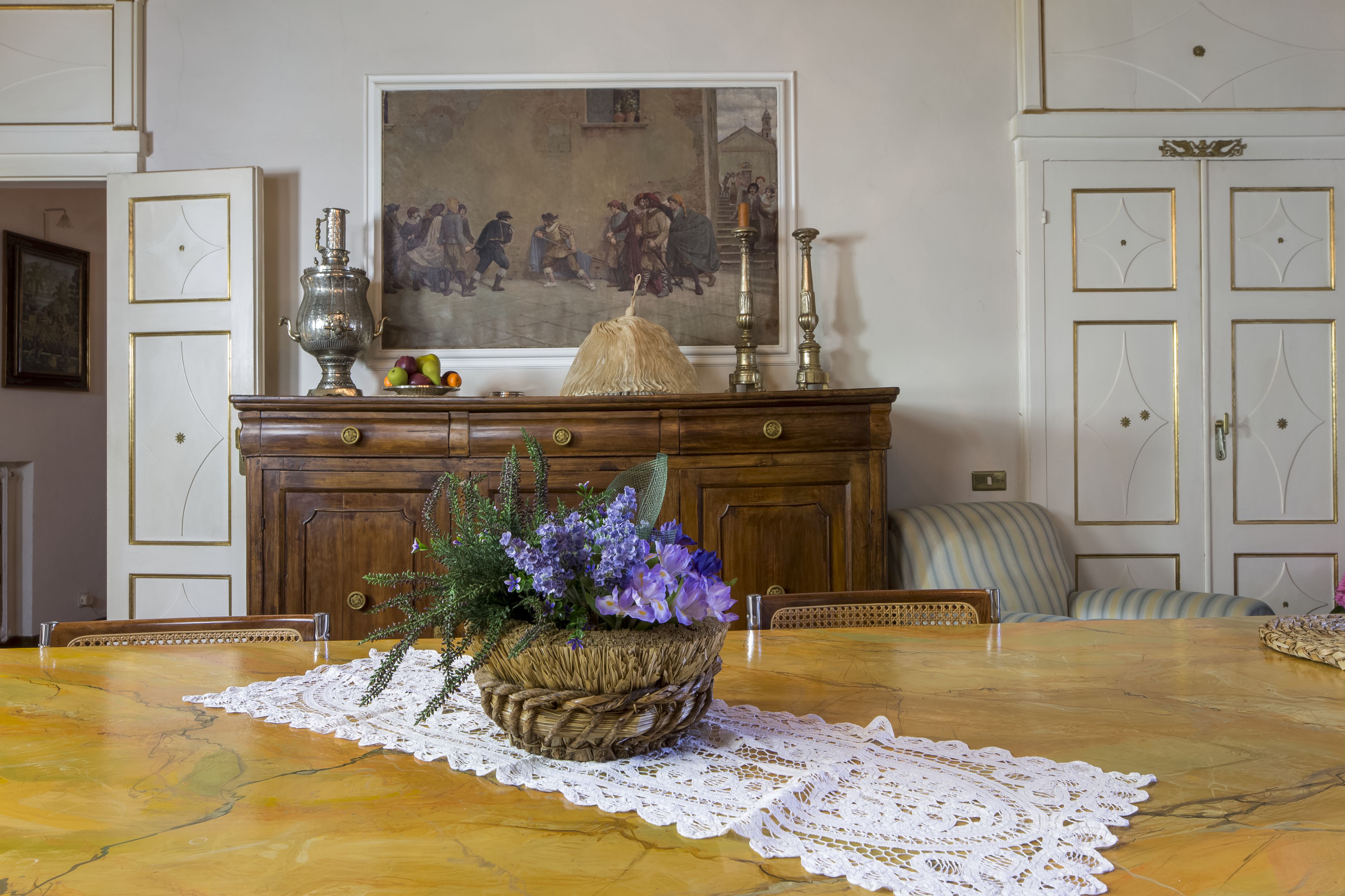 Une table à manger ornée d’un panier tressé rempli de fleurs, d’un chemin de table en dentelle et d’un tableau accroché au mur au-dessus.