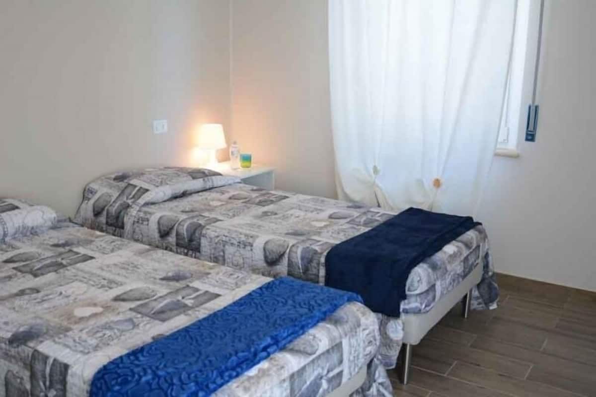 Ein Zimmer mit zwei Betten, jeweils mit einem gemusterten Überwurf und einer blauen Decke. Ein Nachttisch mit Lampe und ein kleiner Tisch mit einer Flasche und einem Behälter sind vorhanden. Das Zimmer verfügt über ein Fenster mit weißen Vorhängen und einen Holzboden.