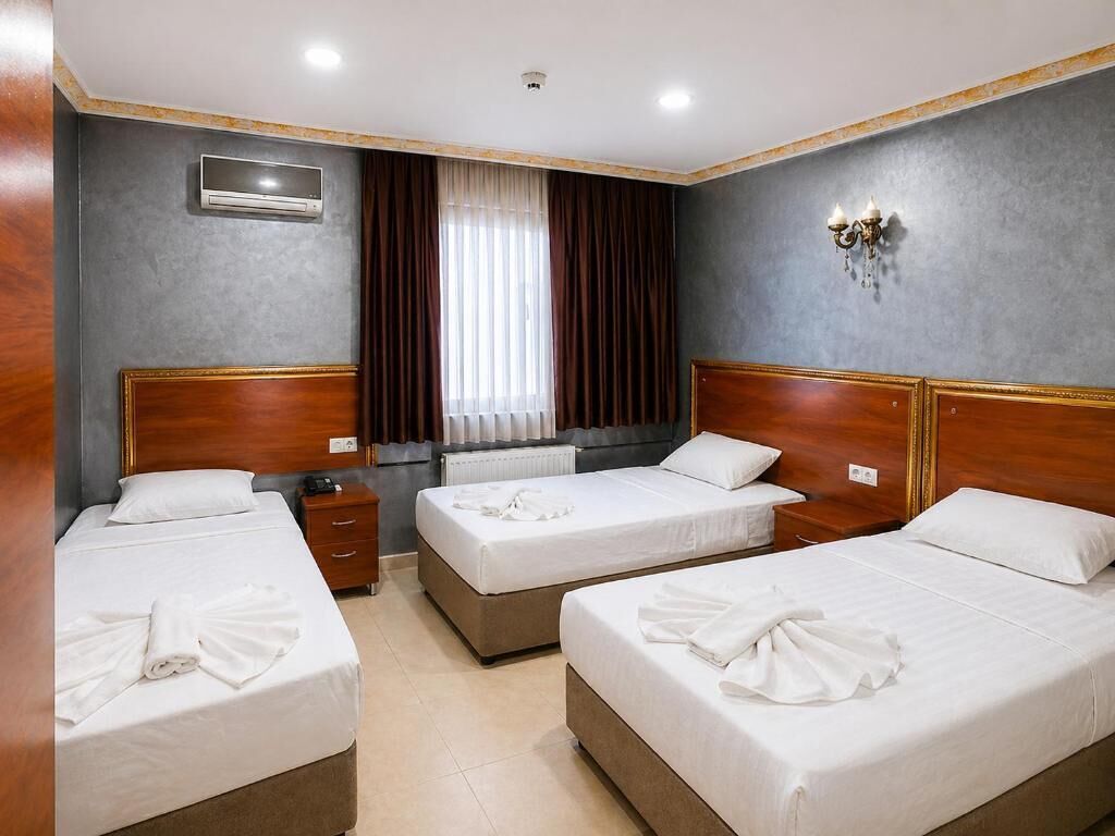A hotel room with three beds, a window with curtains, a wall-mounted air conditioning unit, and a bedside table with a lamp.