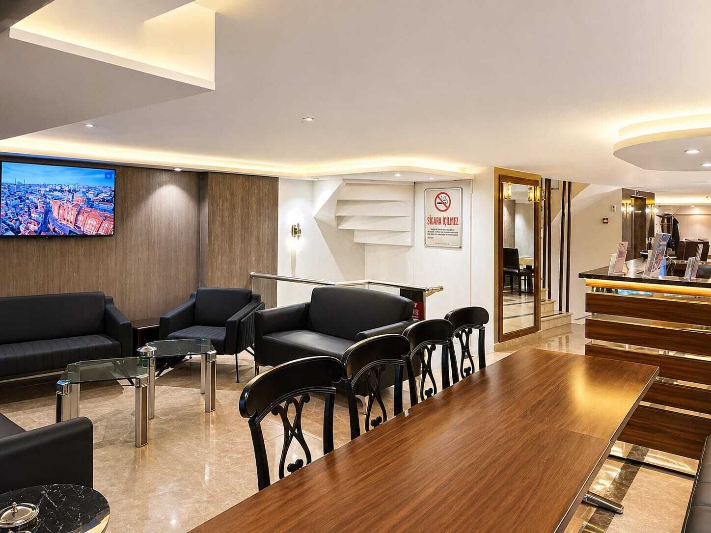 A modern hotel lobby with a reception desk, black leather seating, a large flat-screen TV, and a wooden conference table.