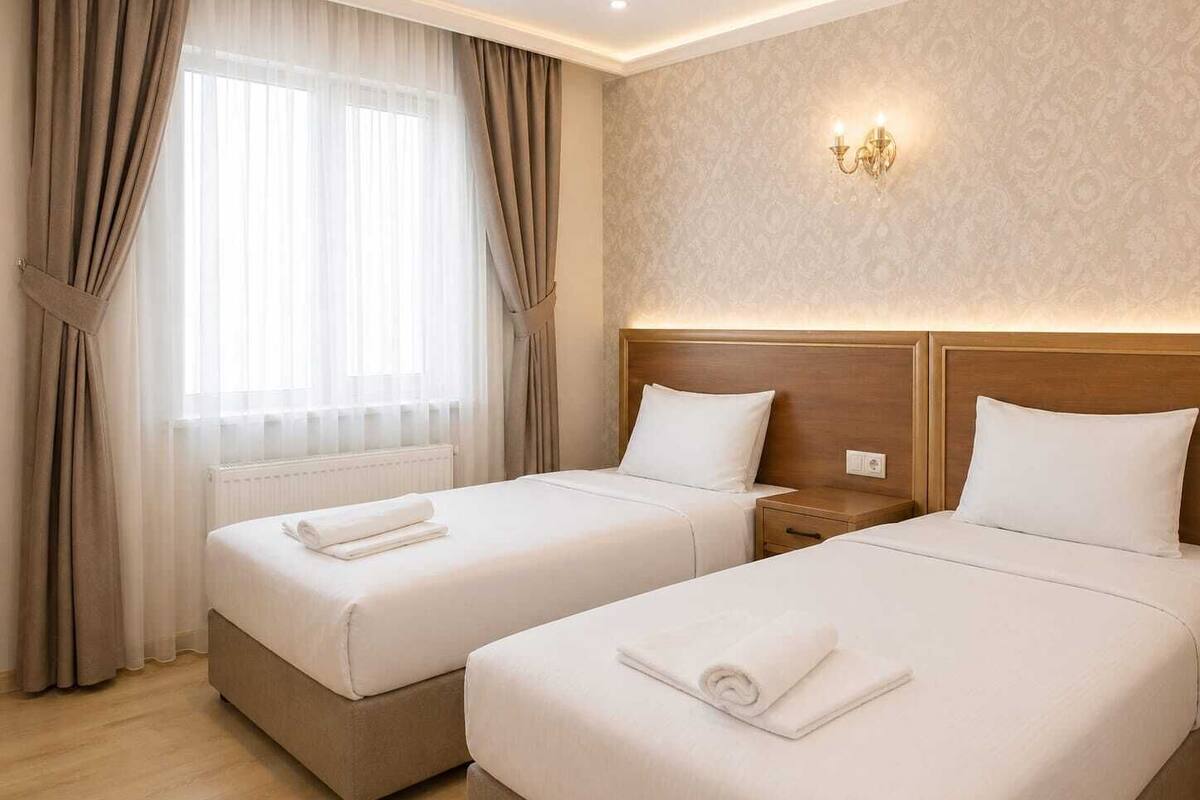 A hotel room with two beds, a nightstand, a wall-mounted light fixture, and a window with curtains.
