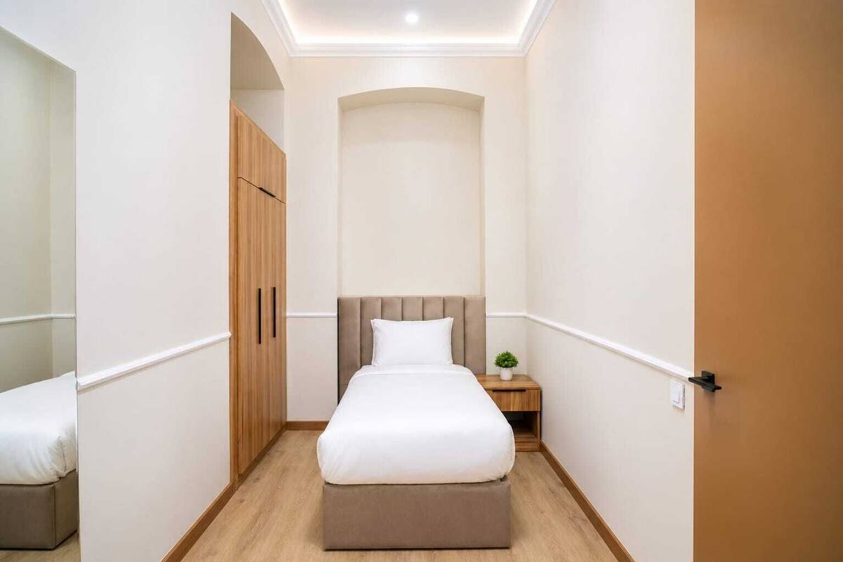 A narrow hotel room with a bed, a wardrobe, a nightstand, and a small plant.