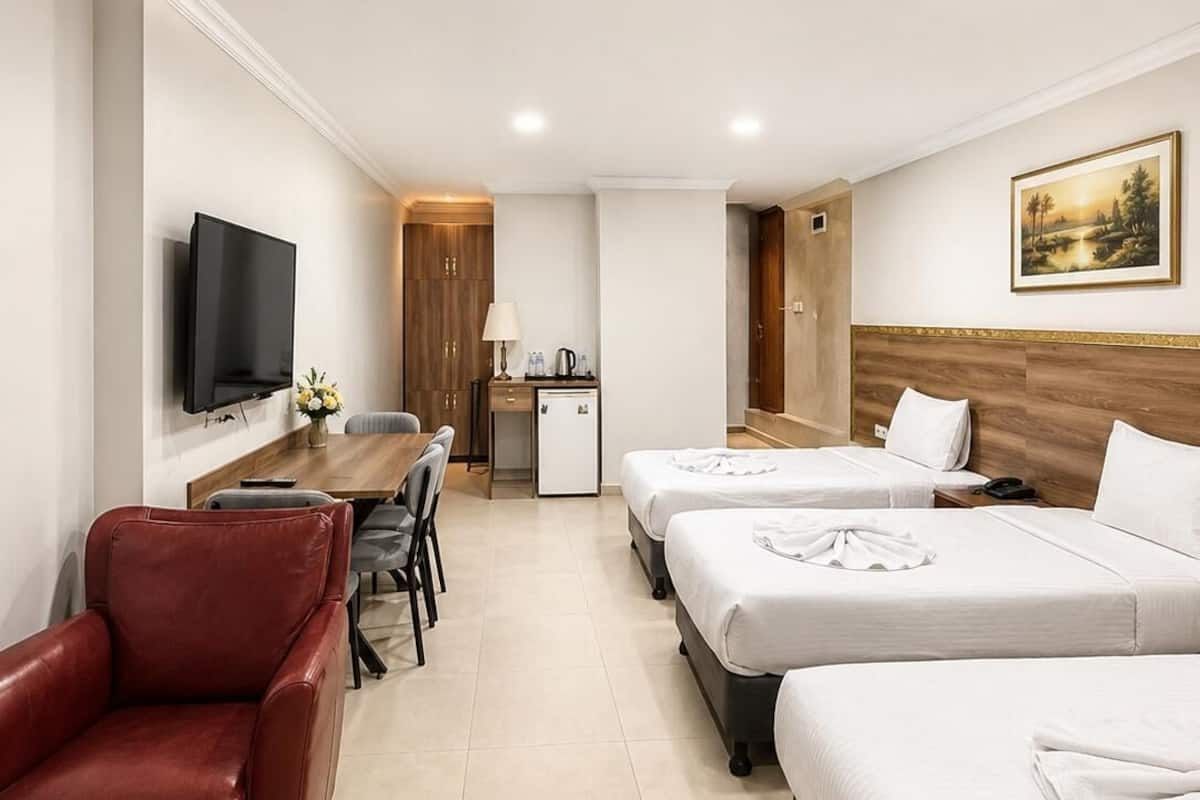 A hotel room with a TV, a desk with a lamp, a small fridge, two beds, a red chair, and a painting on the wall.