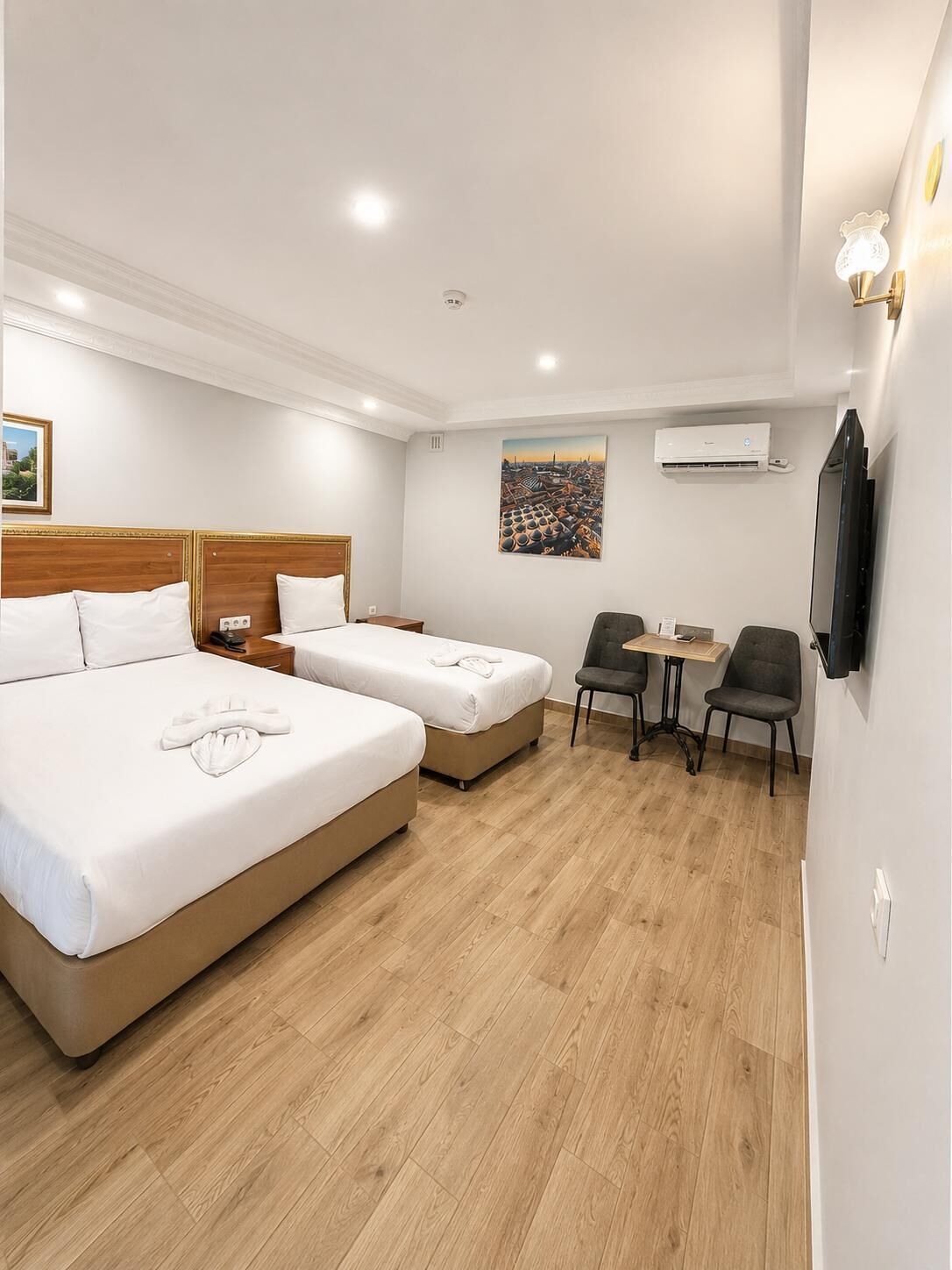 A hotel room with two beds, a small table with chairs, a TV, and an air conditioning unit.