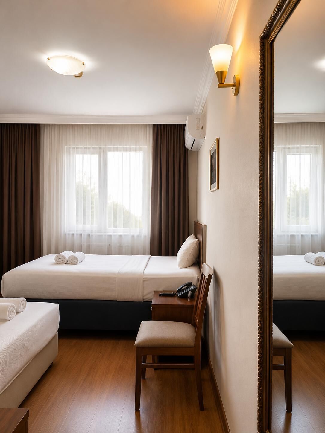 A hotel room with two beds, a desk, a chair, a mirror, and a ceiling light.