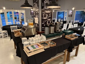 Un buffet de petit-déjeuner est mis en place avec des machines à café, des bouilloires et une variété d’aliments pour le petit-déjeuner disposés sur des tables.