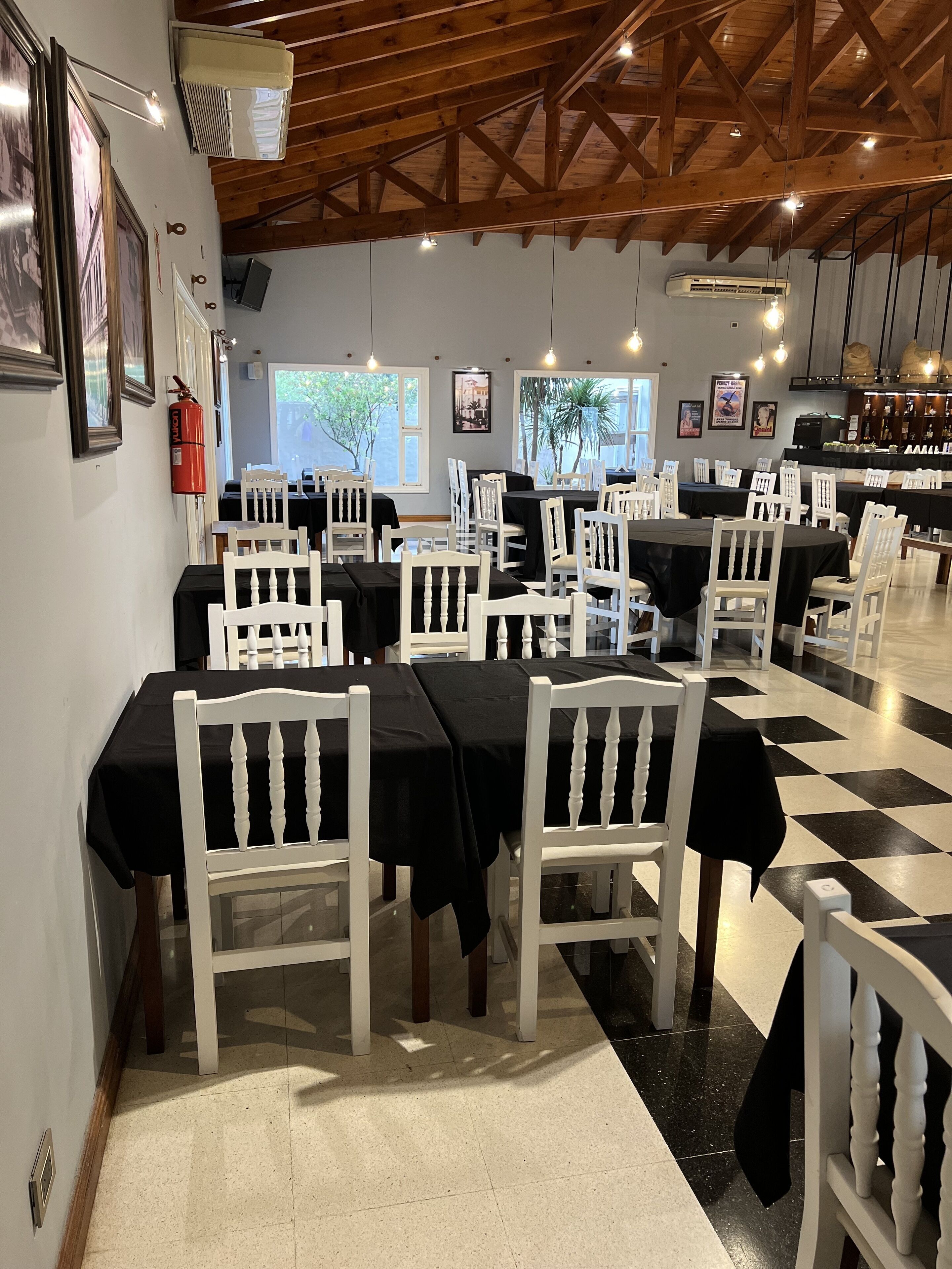 Un restaurante con sillas de madera blanca y manteles negros, un suelo a cuadros y una zona de bar al fondo.