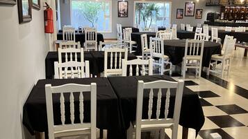 Un restaurante con sillas de madera blanca y manteles negros, un suelo a cuadros y una zona de bar al fondo.
