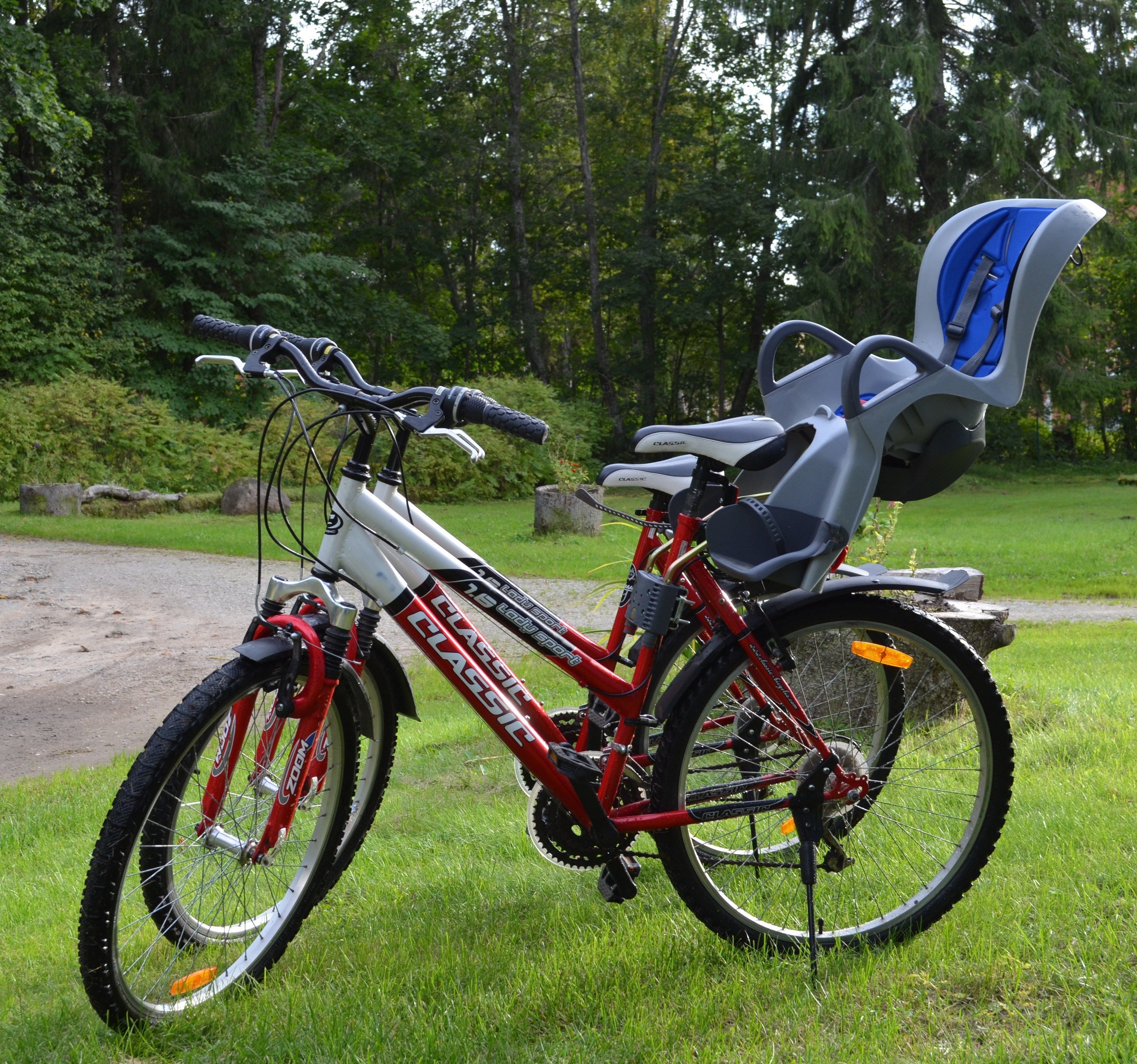 Una bicicleta roja y blanca con un asiento para bebé, estacionada sobre césped con árboles al fondo.