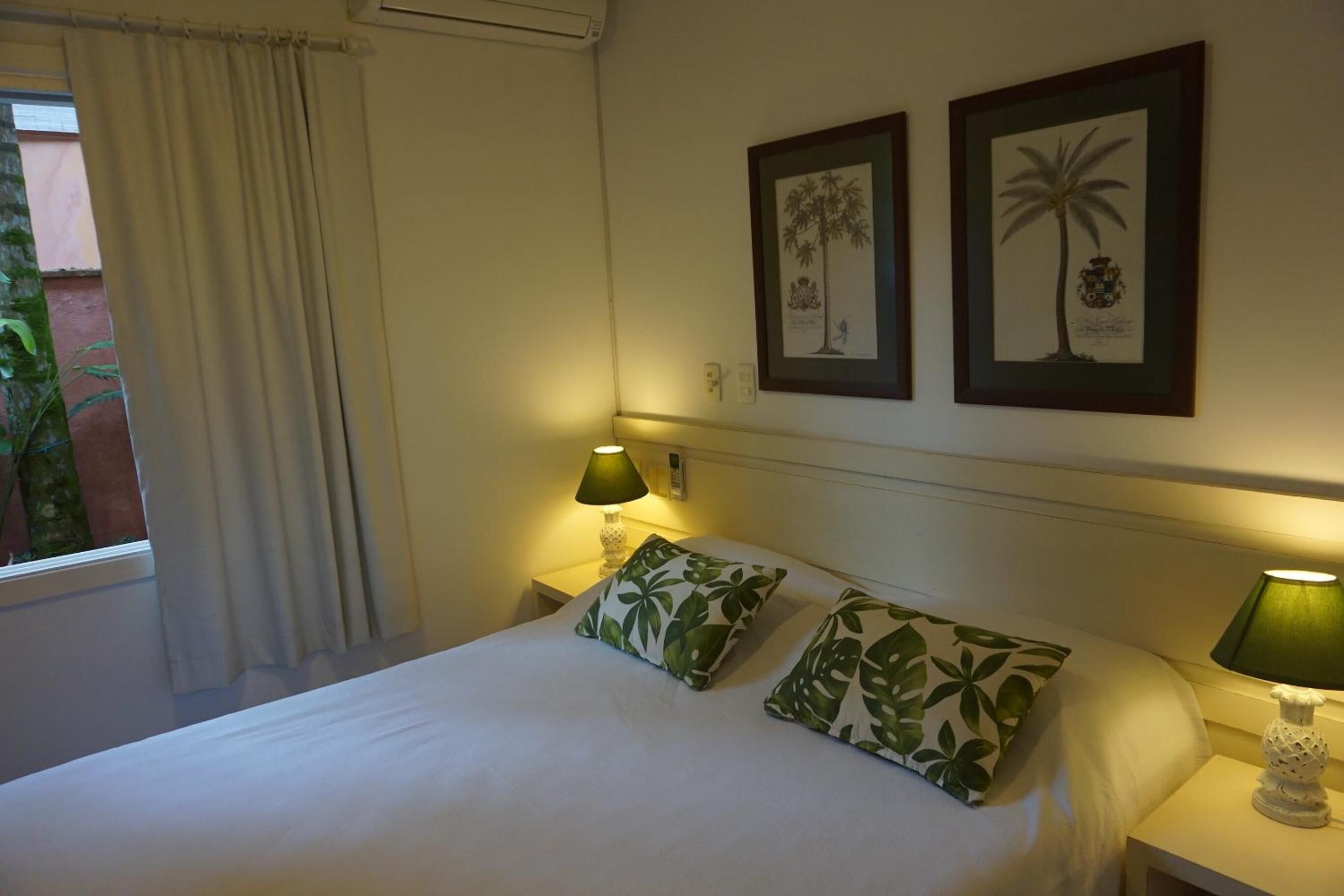 A neatly made bed with two pillows, two wall-mounted lamps, and two framed pictures of palm trees.