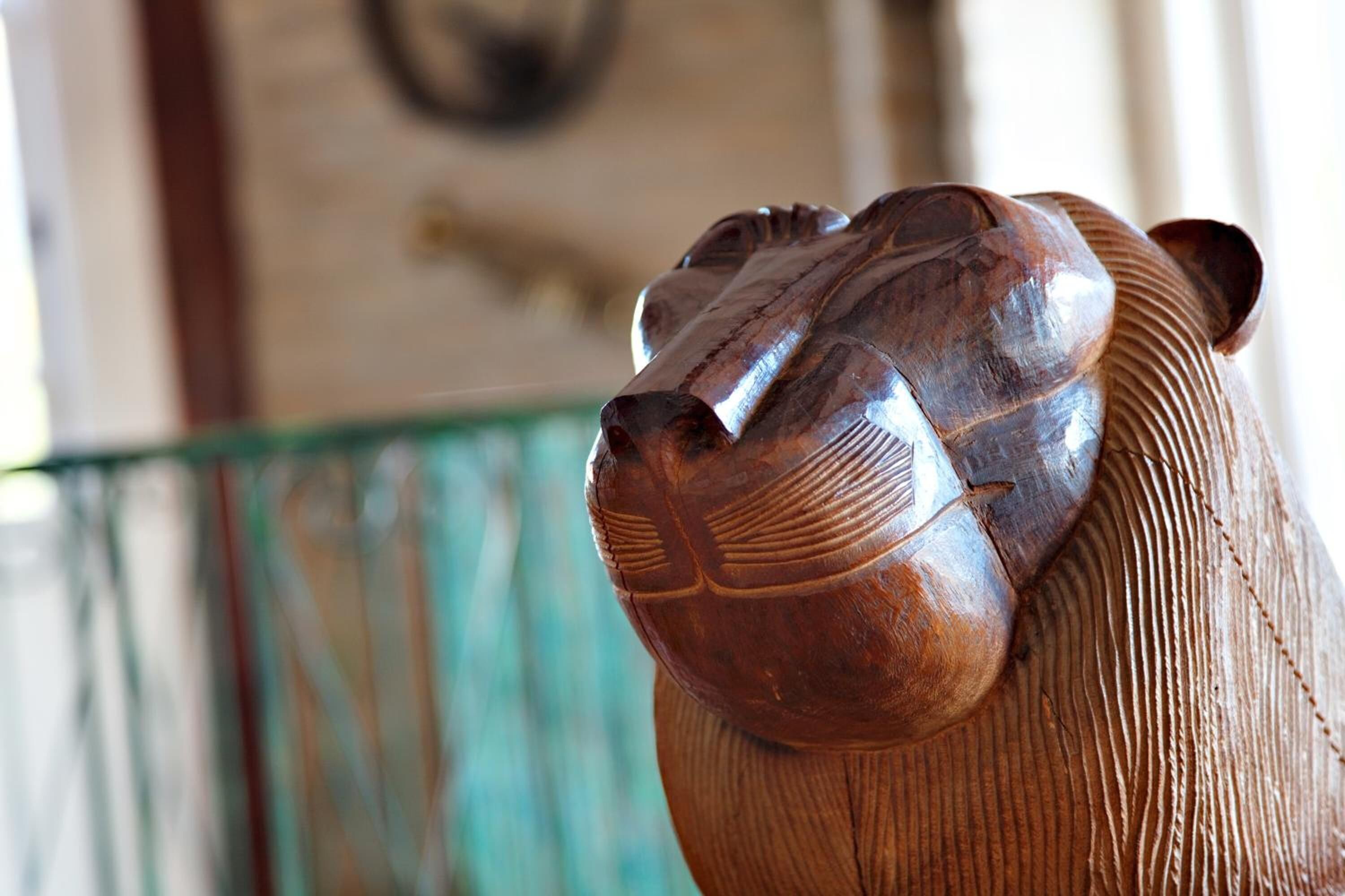 Close-up van een houten beeldhouwwerk met een uitgehouwen gezicht, mogelijk een leeuw of een vergelijkbaar dier, voor een onscherpe achtergrond met een muur en een raam.