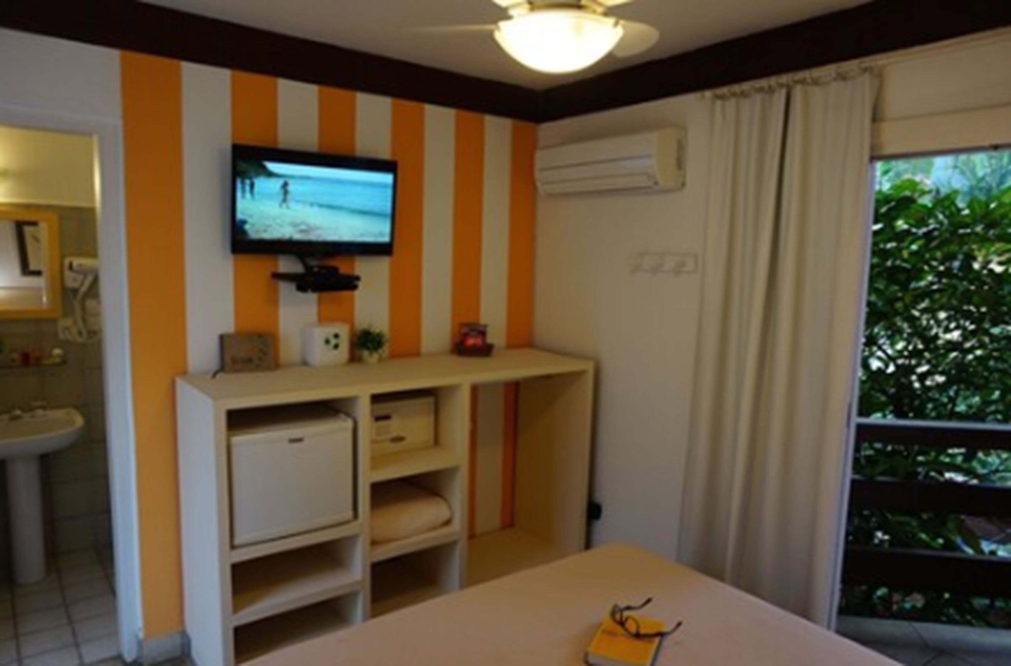 A room with a television mounted on the wall, a bedside table with a book, a window with curtains, and a sink in the corner.