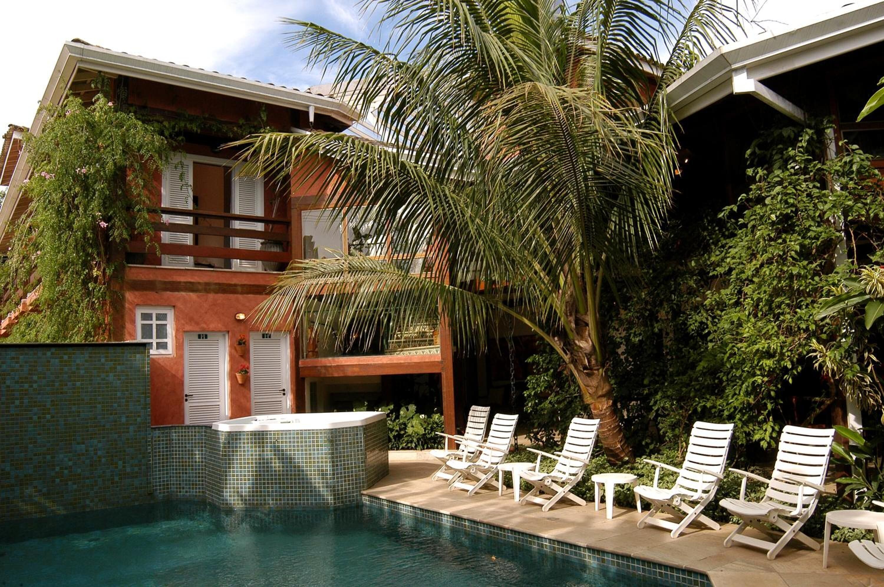 Zona de piscina con sillones de descanso y jacuzzi, rodeada de vegetación exuberante y un edificio con fachada de azulejos.