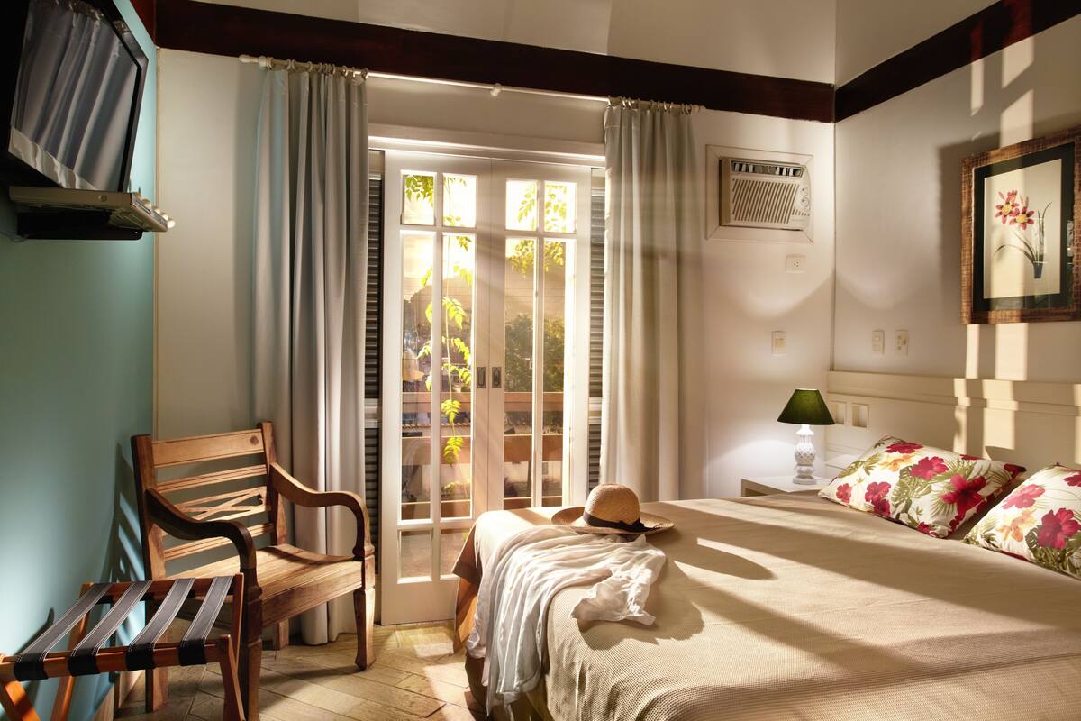 A bedroom with a bed, a wooden chair, a bench, a hat, a lamp, and a window with curtains.