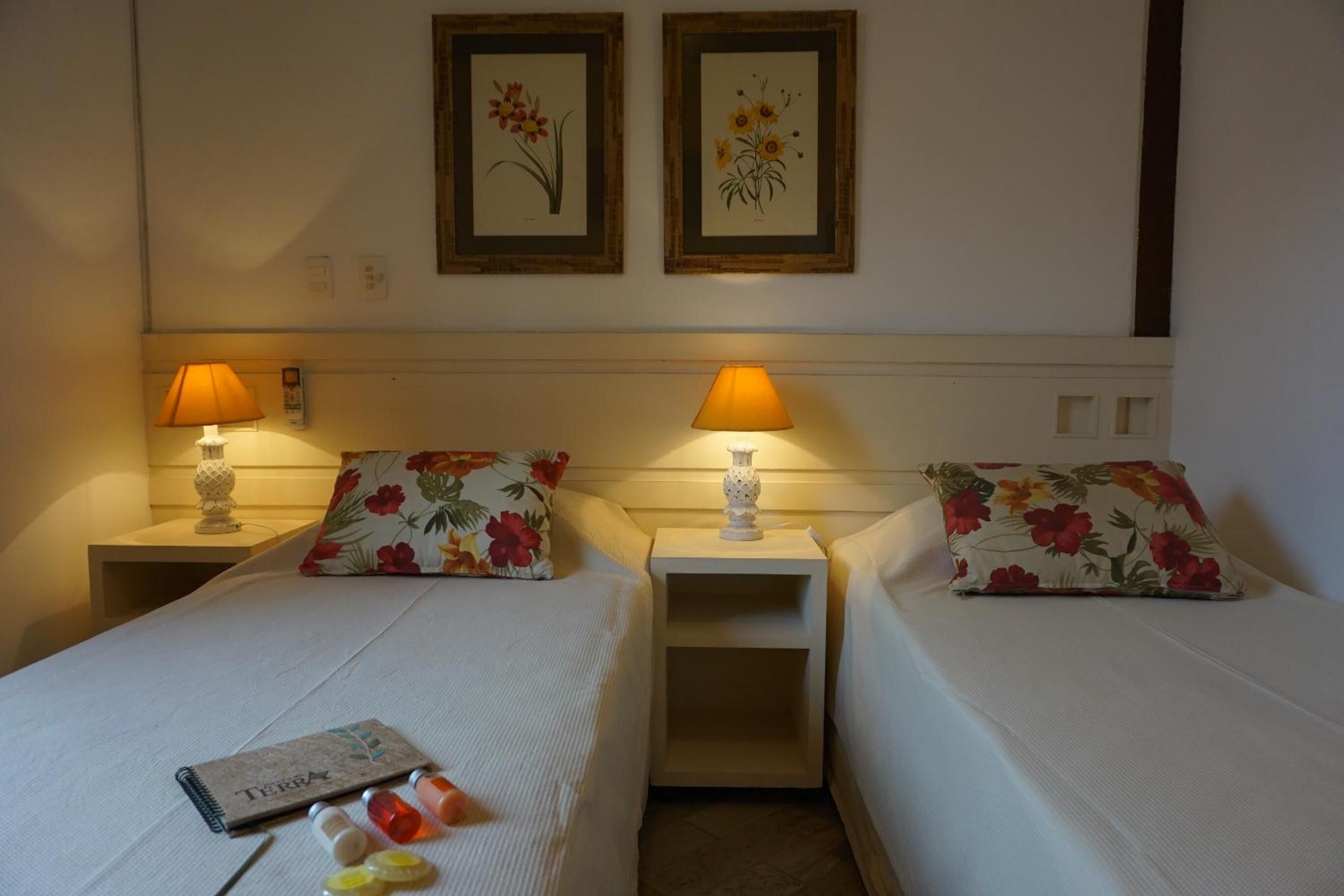 A hotel room with two single beds, each with a floral pillow and a bedside table with a lamp. There are framed botanical prints on the wall above the beds.
