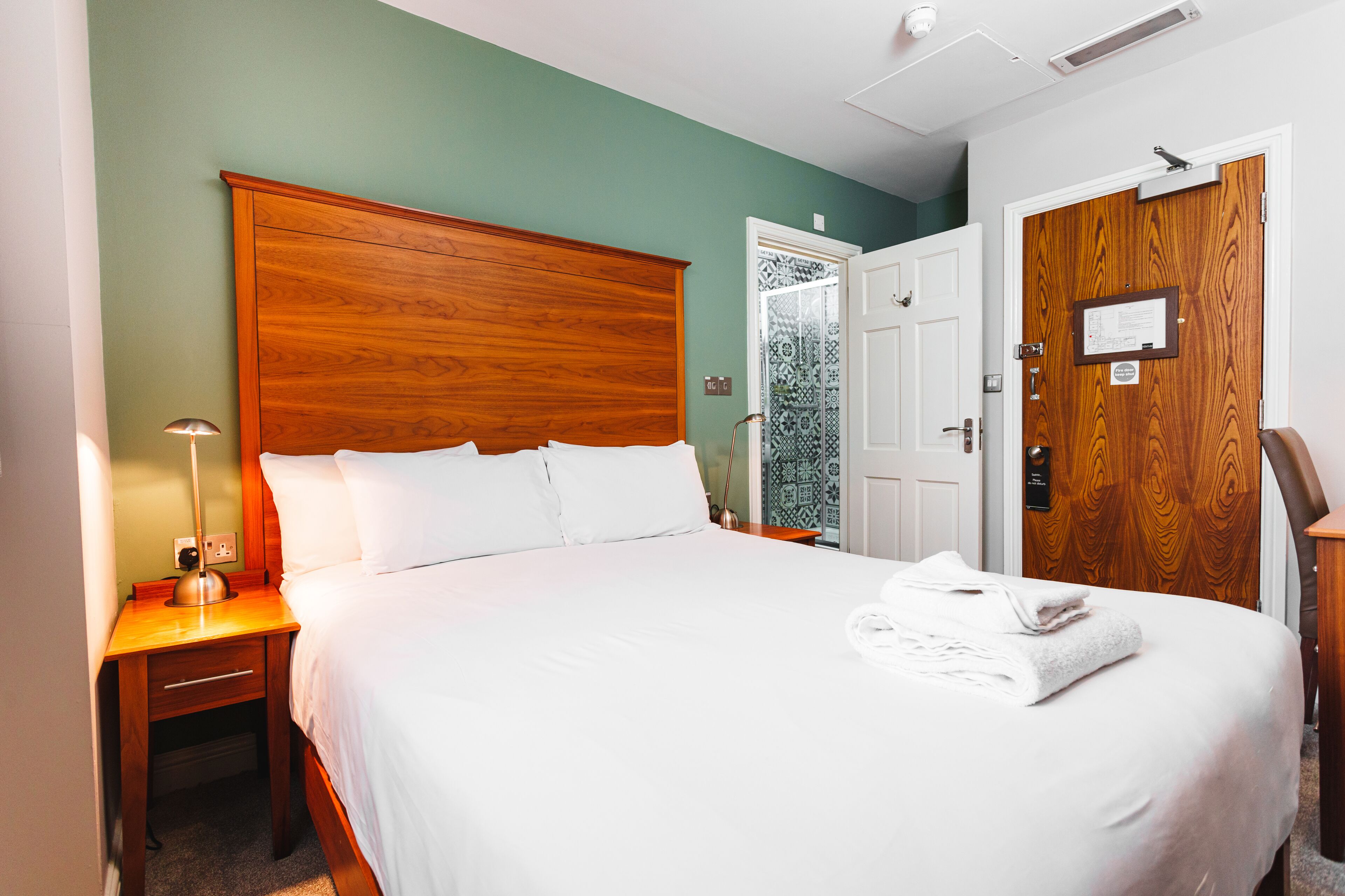 A neatly made bed with white linens and a folded towel on the side, a wooden nightstand with a lamp, and a wooden door with a plaque.