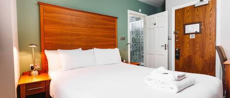 A neatly made bed with white linens and a folded towel on the side, a wooden nightstand with a lamp, and a wooden door with a plaque.