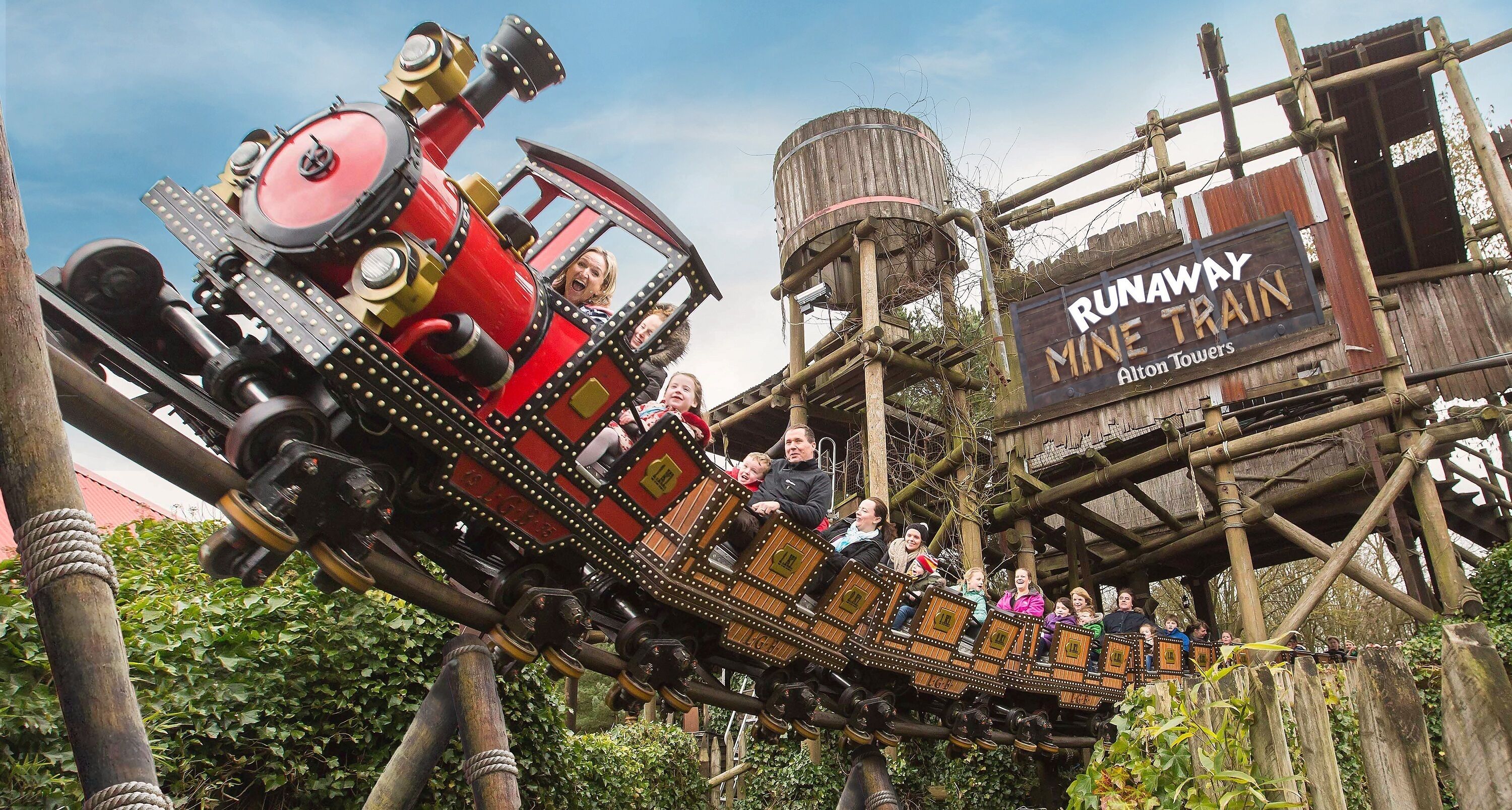 A wooden roller coaster with passengers, surrounded by a rustic wooden structure and greenery.