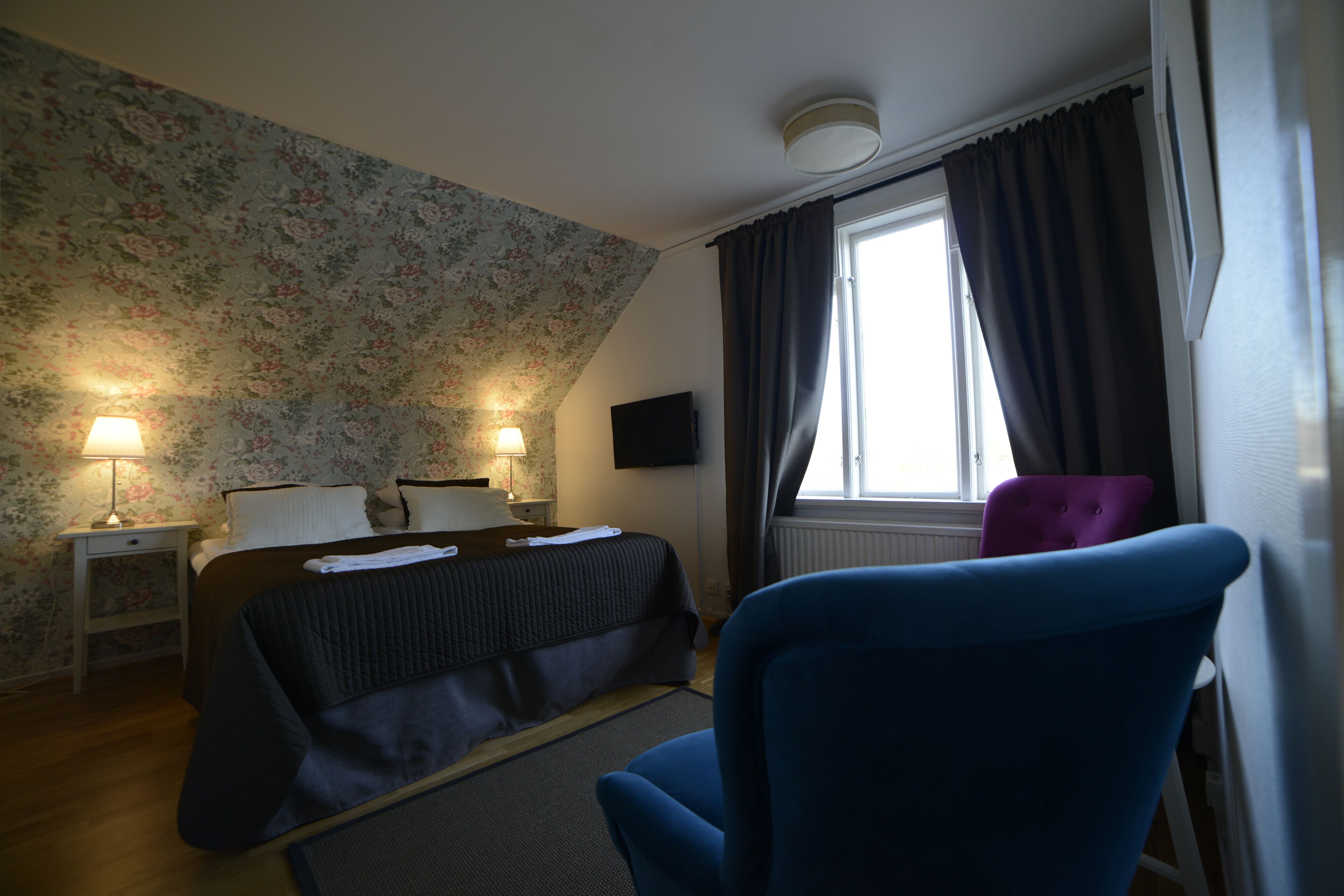 Une chambre à coucher avec un lit, deux lampes de chevet, un téléviseur à écran plat, une fenêtre avec des rideaux et un fauteuil bleu.