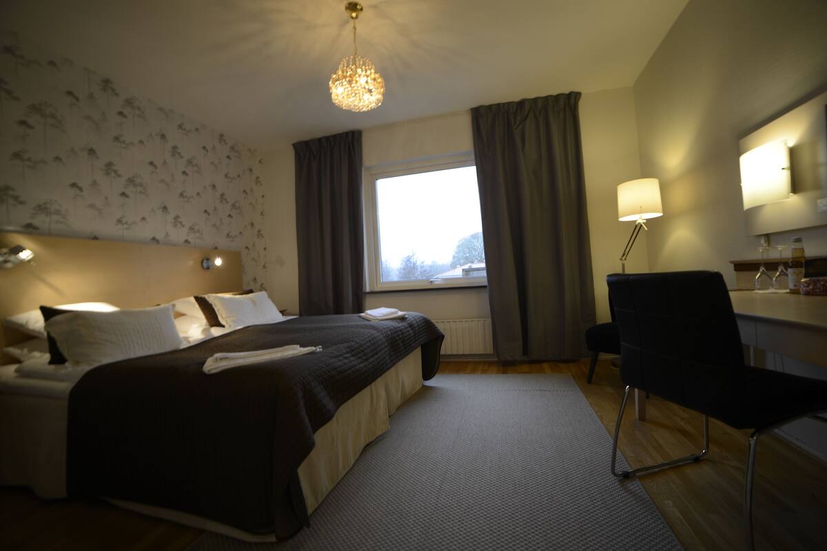 Une chambre d’hôtel avec un lit, un bureau, une chaise, une lampe et une fenêtre avec des rideaux.