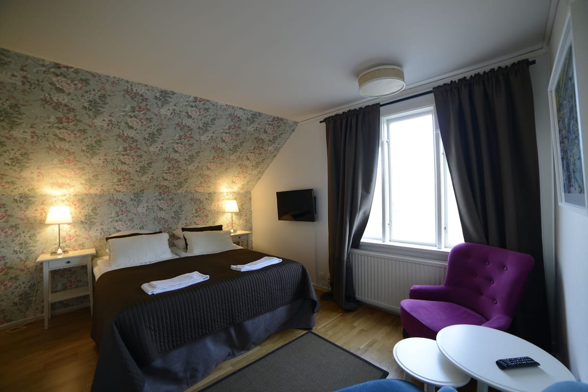 Une chambre d’hôtel avec un lit, une chaise, une petite table, un téléviseur et une fenêtre avec des rideaux.