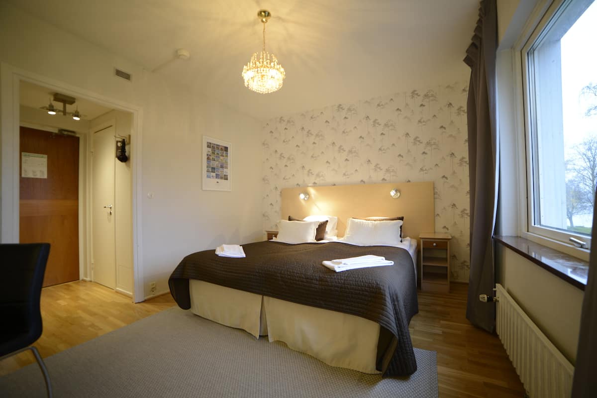 Une chambre d’hôtel avec un grand lit, une table de chevet, une chaise, une fenêtre avec des rideaux et un lustre.