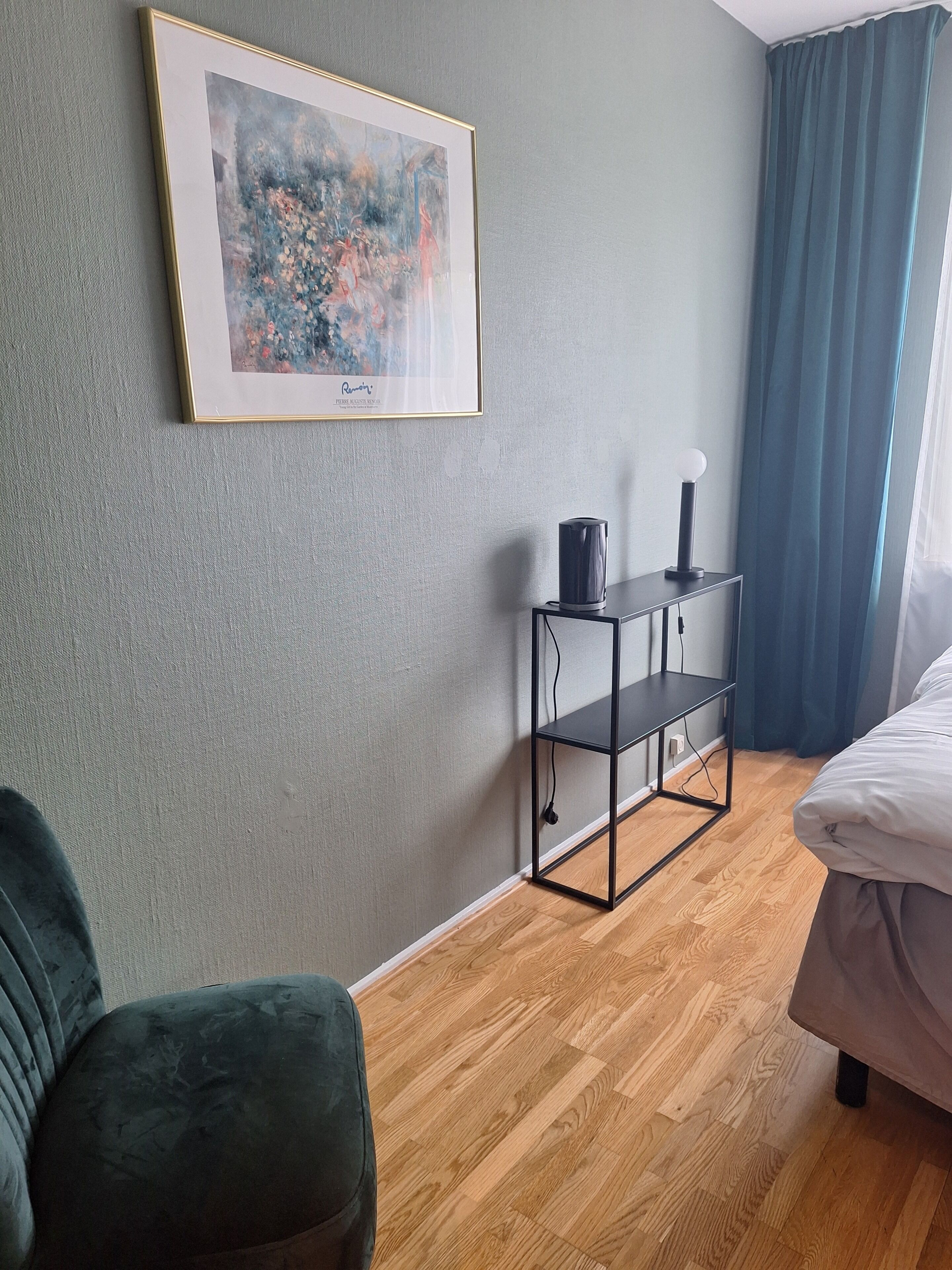 Une pièce avec un tableau encadré, une étagère noire sur laquelle se trouve une lampe, un fauteuil vert et un lit.