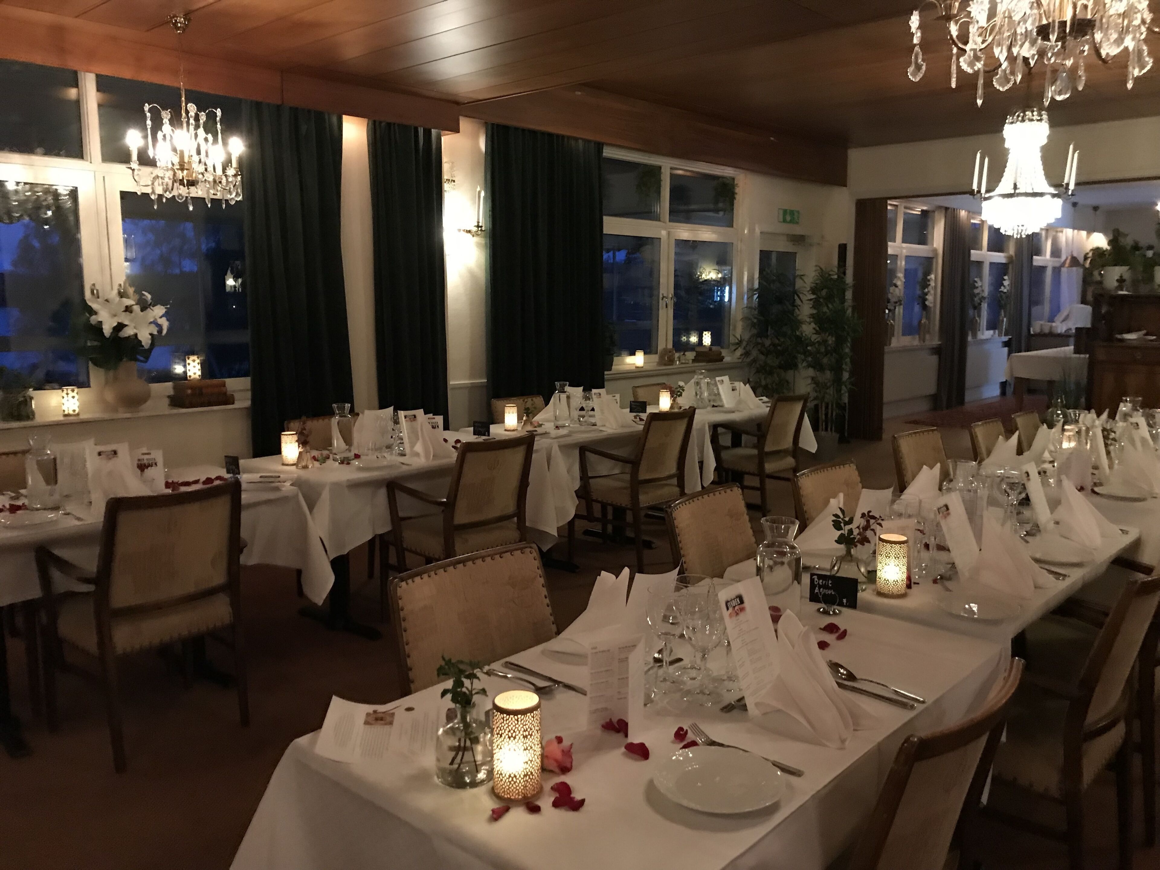 A restaurant with multiple tables set for dining, featuring chandeliers, candles, and a view of the night sky through large windows.