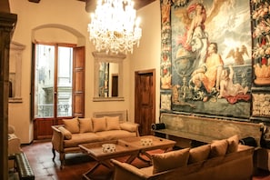 Un vaste salon agrémenté d’un lustre, d’une grande tapisserie et offrant une vue sur la ville par la porte ouverte.