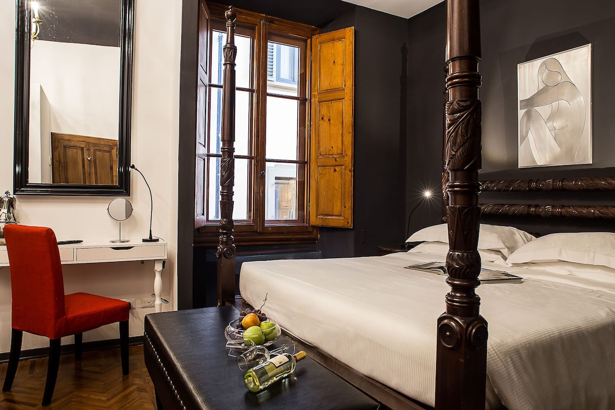 A bedroom with a four-poster bed, a desk with a chair, a window with wooden shutters, and a painting on the wall.