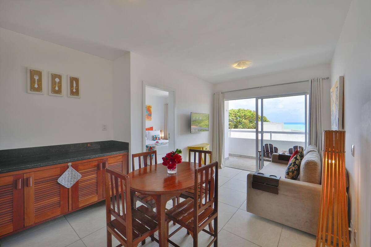 A modern kitchen with wooden cabinets, a dining table with chairs, a small island with a TV, and a balcony with a sofa and a view of the sea.