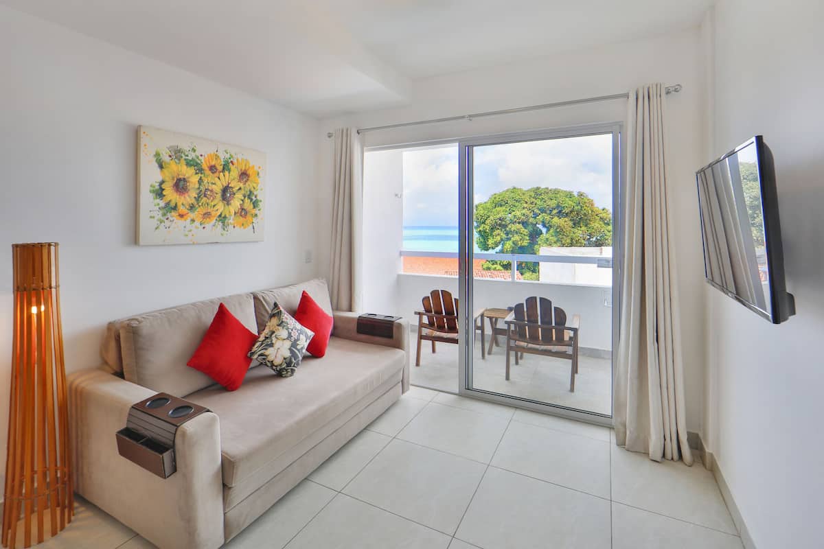 A modern living room with a sofa, red and floral cushions, a painting of sunflowers, a TV, and a balcony with a view of the ocean.