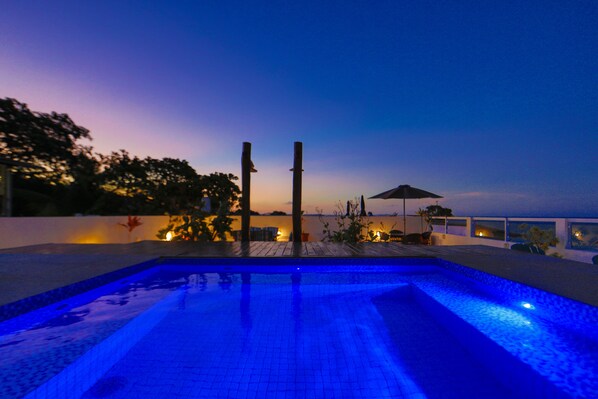A swimming pool at dusk with a sunset view, surrounded by plants and a wooden deck.