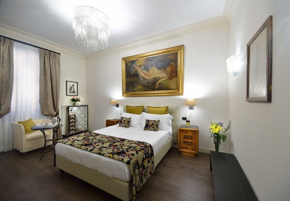 A hotel room with a large bed, a sitting area with a chair and a small table, a painting on the wall, and a bench along the wall. - Suites Rome Condotti (Rome)