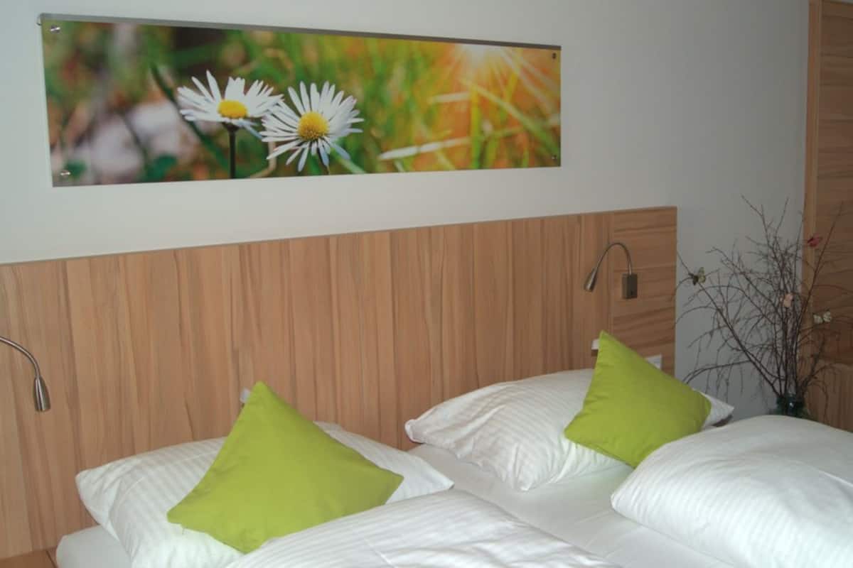 A bedroom with two beds, each with white bedding and a green pillow. There is a wooden headboard, a wall-mounted light, and a framed picture of flowers above the beds.