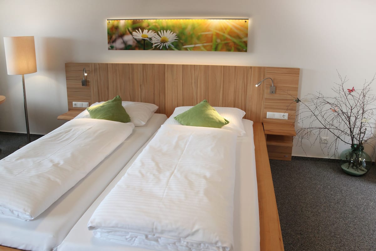 A double bed with white linens and green pillows, a wooden headboard, a wall-mounted TV, and a vase with branches on the right.