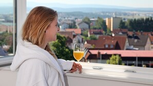 Iemand zit bij een raam met een glas sinaasappelsap, met uitzicht op een stad met rode daken en groen.