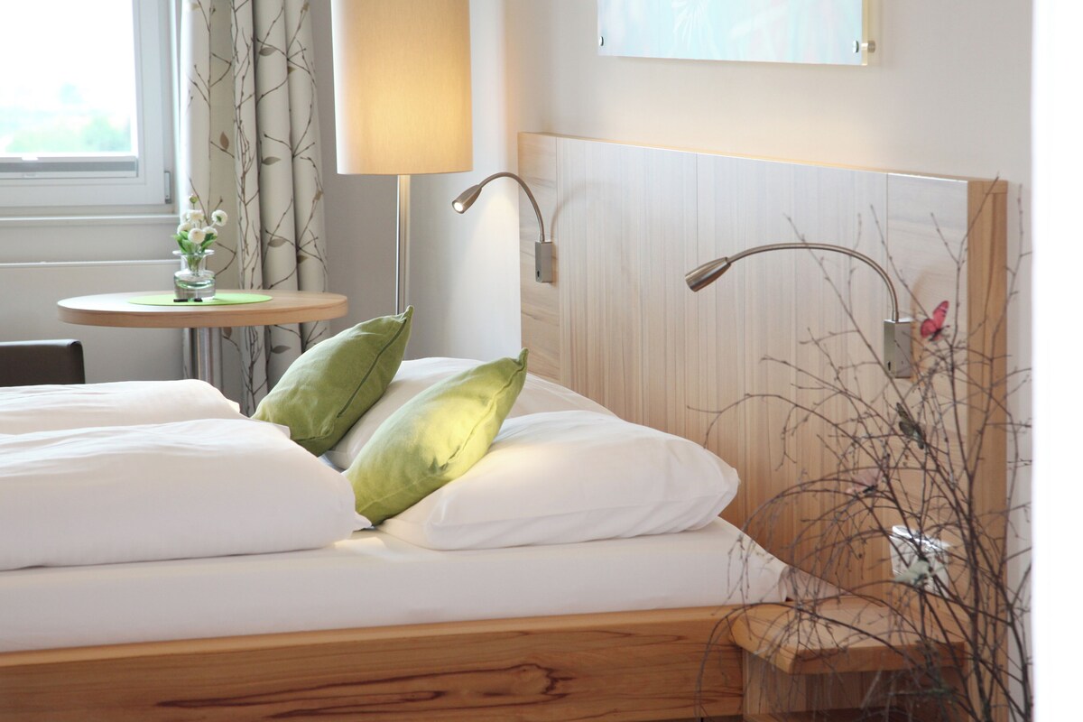 Een hotelkamer met een houten bed, een klein rond tafeltje met een bloevas en een lamp.