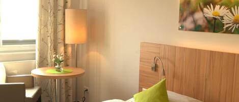 Una habitación de hotel con cama, una mesita de noche con lámpara, una silla y un cuadro de flores en la pared.