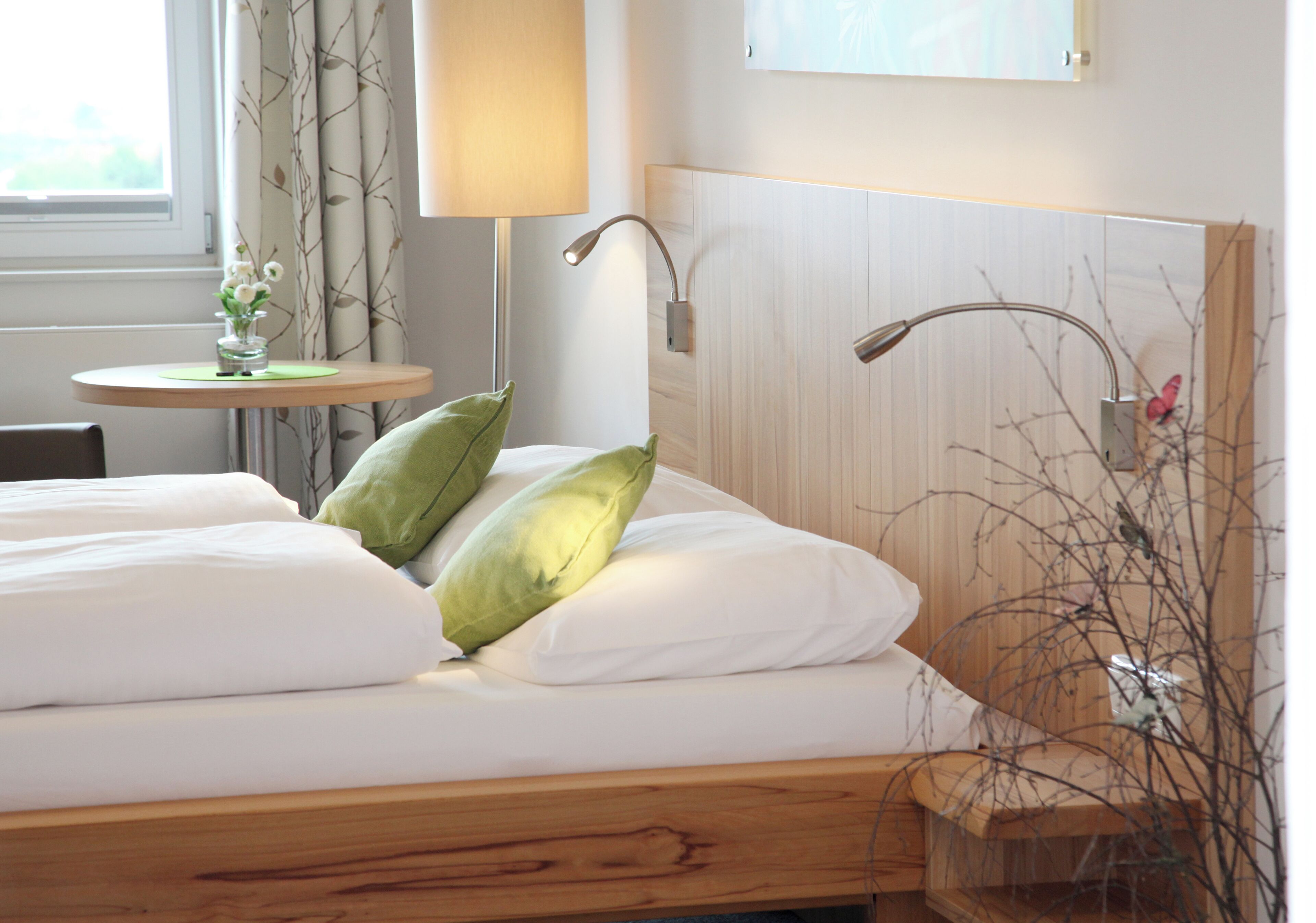 Een hotelkamer met een houten bed, een klein rond tafeltje met een bloevas en een lamp.