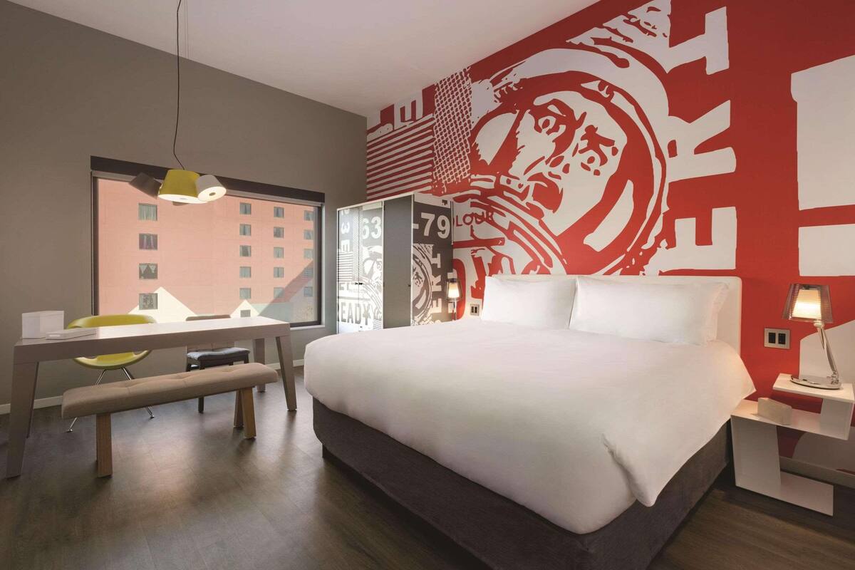 A hotel room with a large bed, a desk with a chair, a bench, a bedside table with a lamp, and a wall with large red and white artwork.