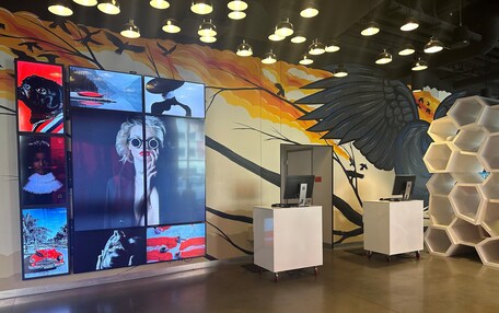 A modern interior with a wall-mounted digital display, white desks, and hexagonal shelving units.