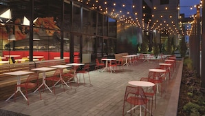 An outdoor patio area with tables and chairs, string lights overhead, and a view of a building with large windows. - Radisson RED Minneapolis Downtown (Minneapolis)