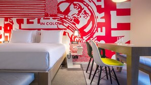 Habitación de hotel con cama, escritorio y silla. Una pared presenta un diseño rojo y blanco.