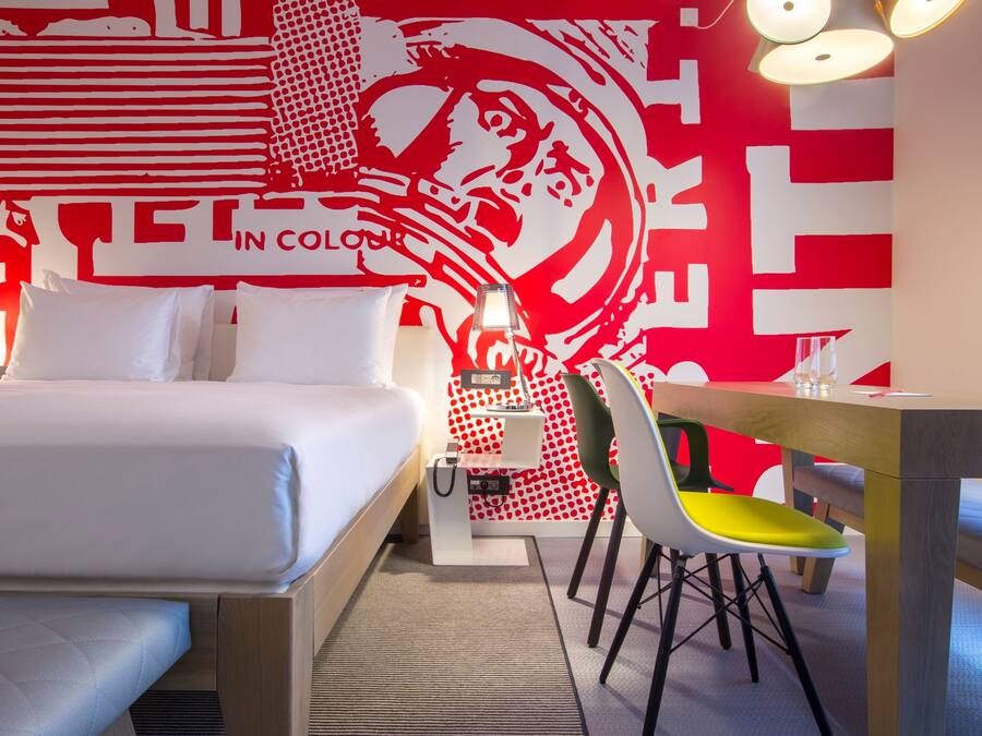 A hotel room with a bed, a desk, and a chair. There is a wall with a red and white design.