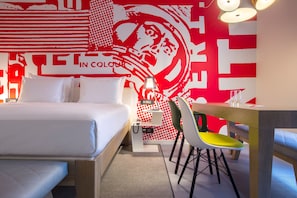 Habitación de hotel con cama, escritorio y silla. Una pared presenta un diseño rojo y blanco.